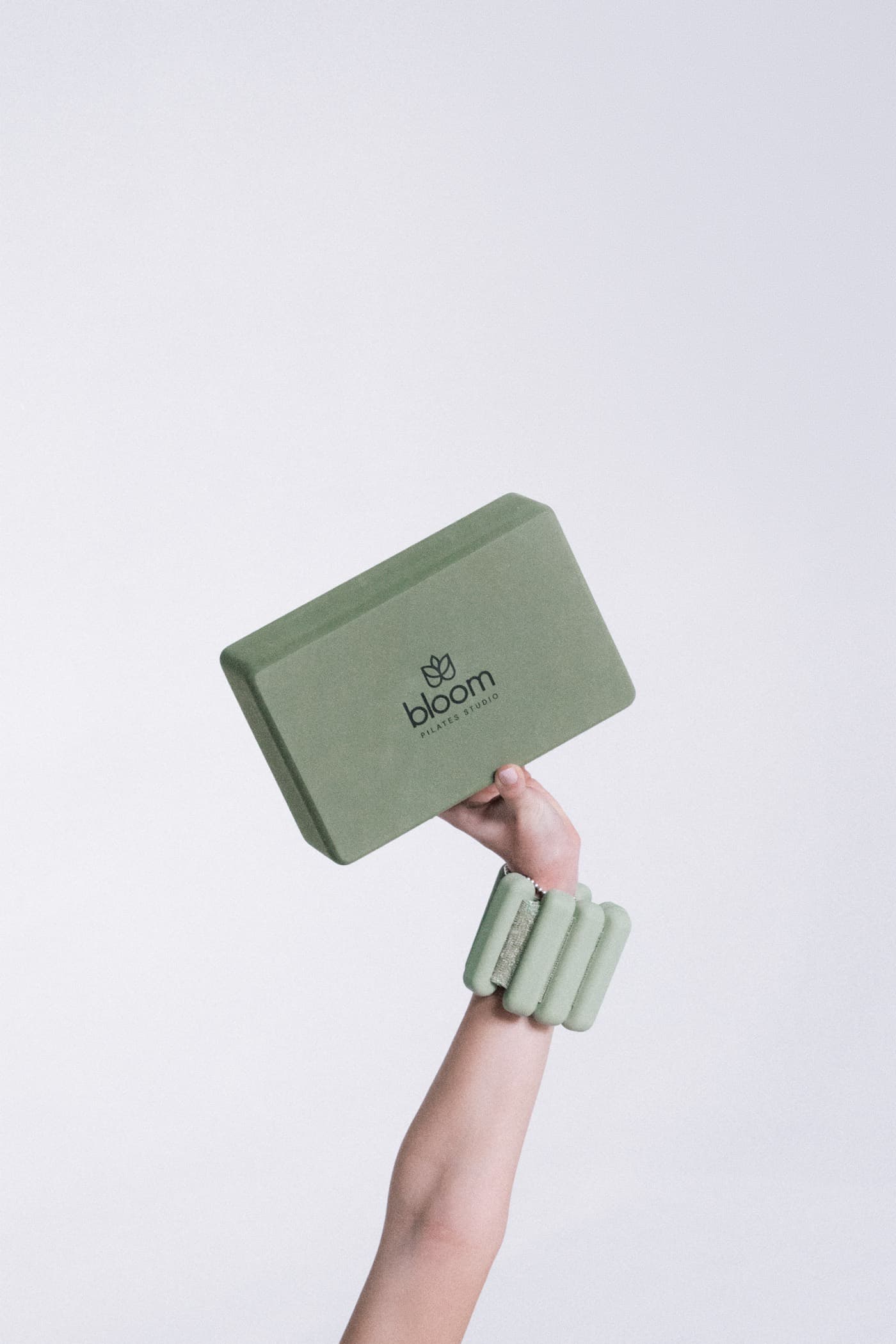 Mano en alto sosteniendo un bloque verde con el logo de Bloom, con pesa de muñeca sage